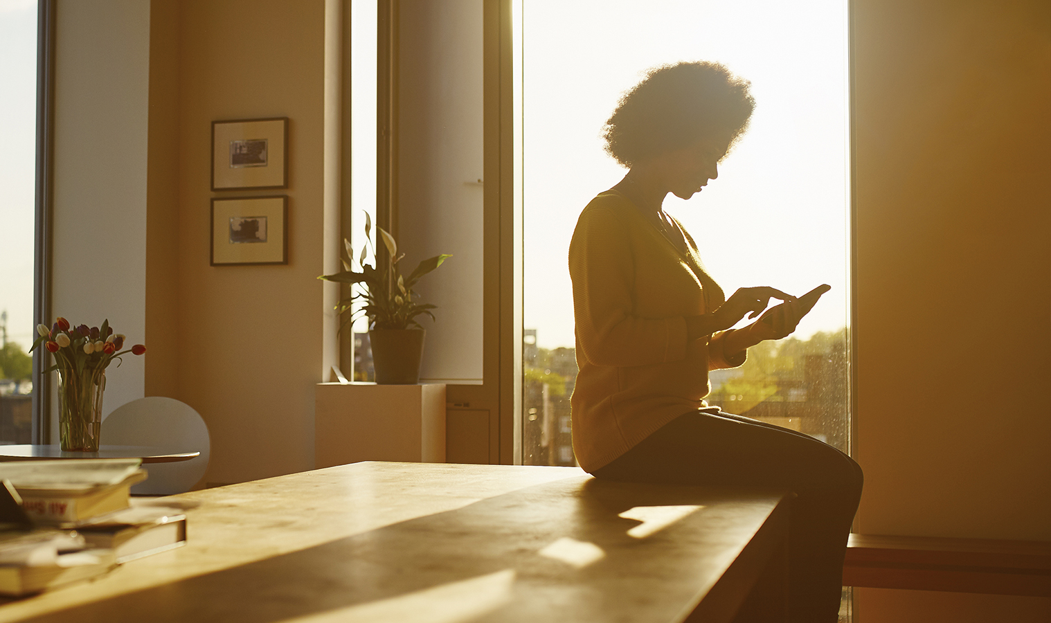 A silhouette of a woman sitting on her kitchen table on  her phone with a sunset behind her