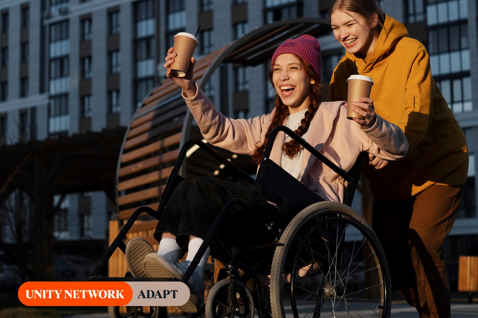 two young women, one on a wheelchair holding 2 cups of cofee and her friend pushing her on