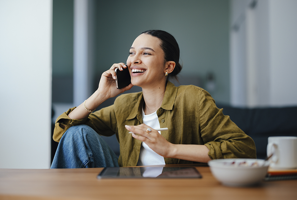 Woman smiling on phone with stylus in hand and tablet on desk infront of her