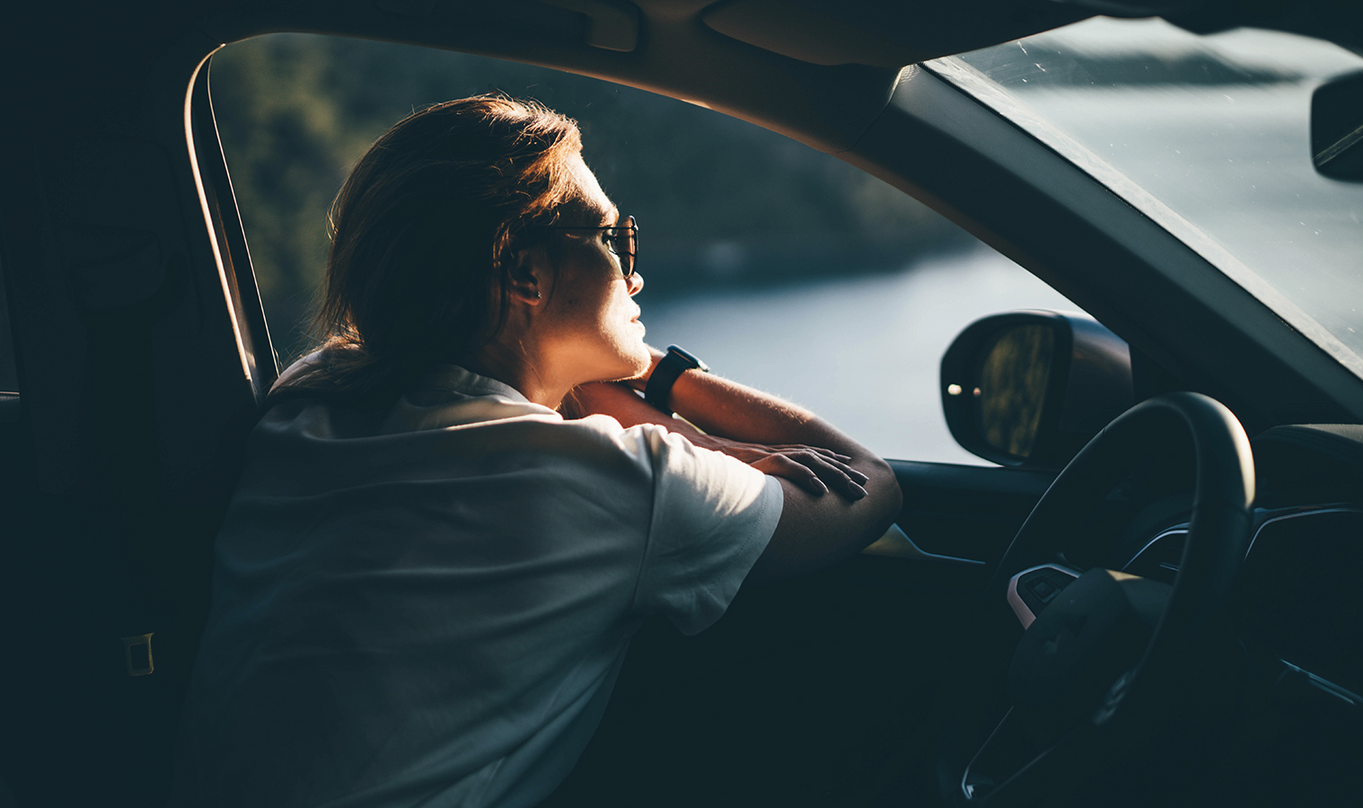 woman sitting in a car in drivers seat, looking out an open window while resting her head on her arms