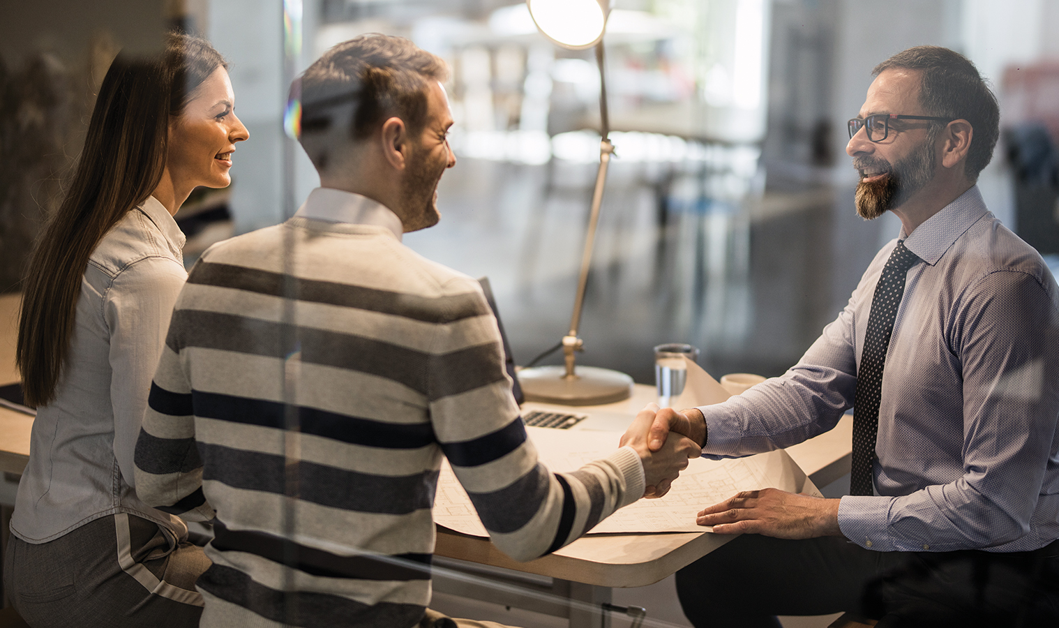 meeting in an office of a couple with another proffessional, men are shaking hands over a desk with house blueprints 