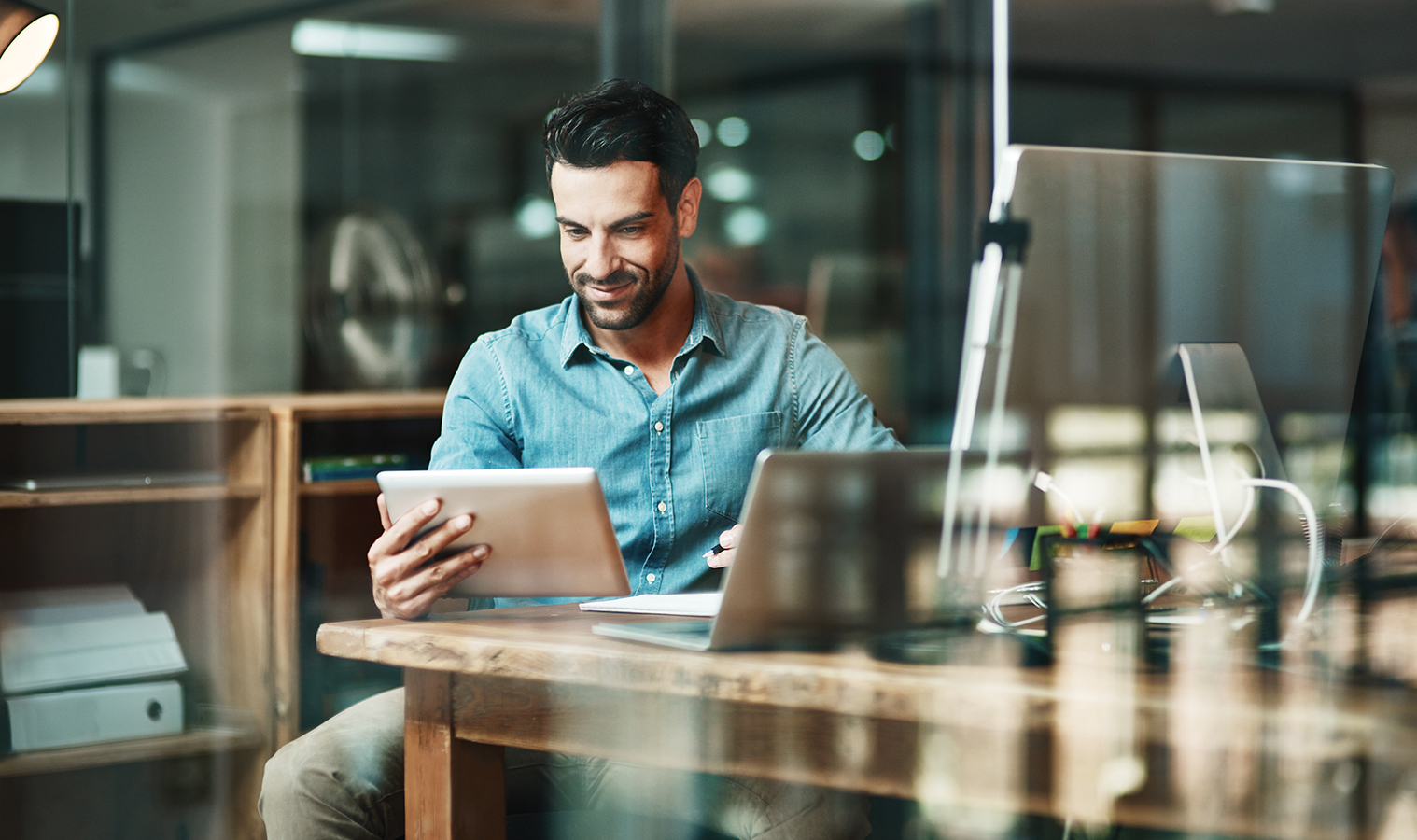 man sitting in an office at a desk with monitors, laptop and holding a tablet in hand