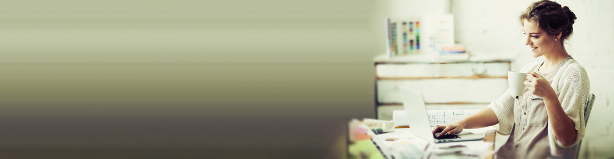 A woman in a knitted jumper holding a cup of coffee is sitting at a table while looking at her laptop