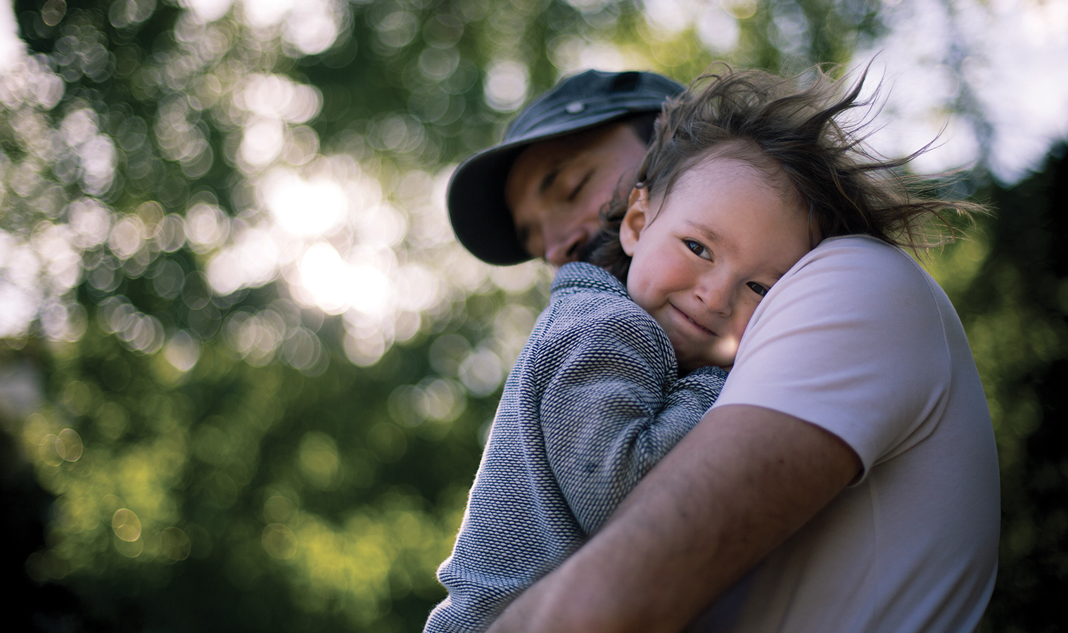 a man holding a smiling toddler cuddling to his shoulder, trees in a background