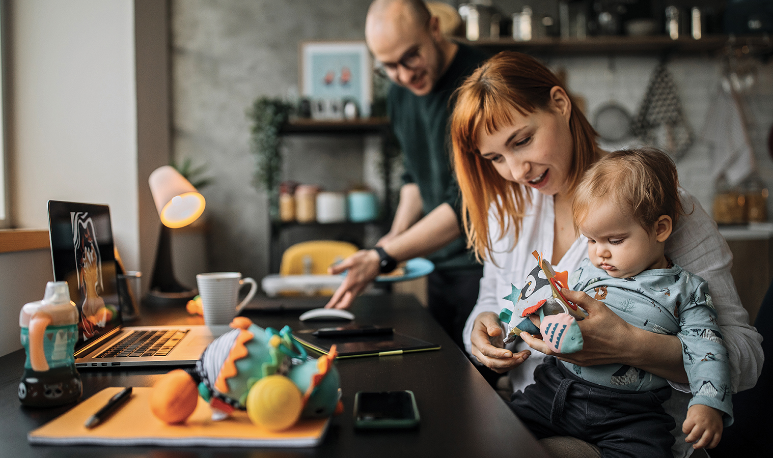 yourg family with a toddler sitting at a kitchen table, father is placing food , mom is playing with the baby 