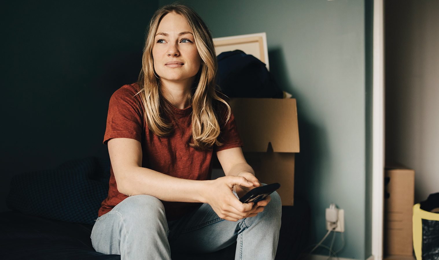 A blonde hair woman in a red tshirt sitting on cardboard boxes  with her phone in her hands
